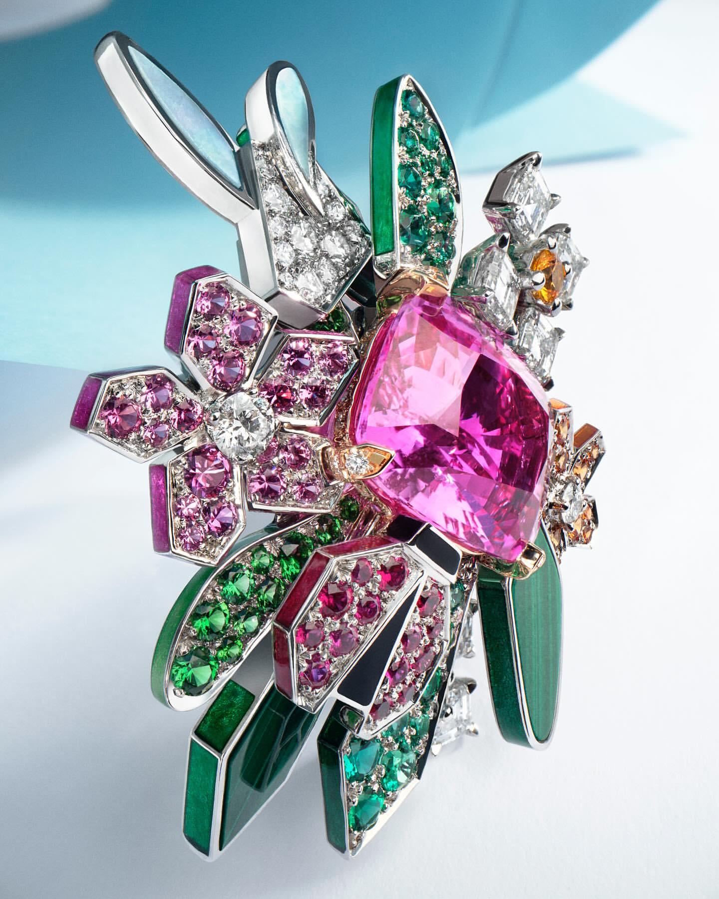 Close-up studio shot of a floral brooch with gems of pink, green, white, and orange, set in silver metal on a gradient white and blue background.