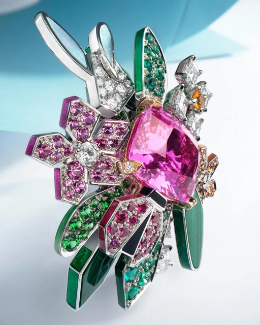 Close-up studio shot of a floral brooch with gems of pink, green, white, and orange, set in silver metal on a gradient white and blue background.