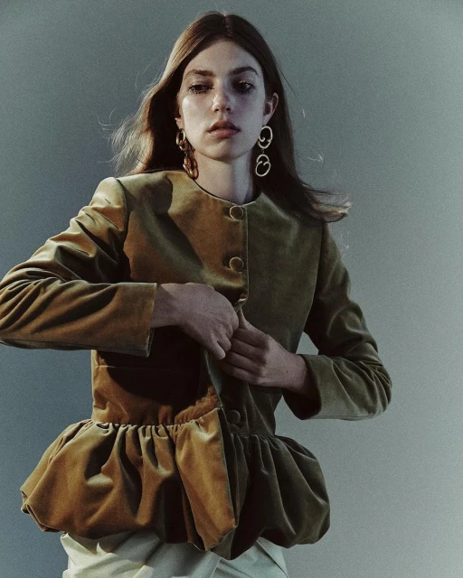 A young adult female buttons a short, fitted, light-brown velvet jacket in a studio setting with a grey background and soft, diffused lighting.