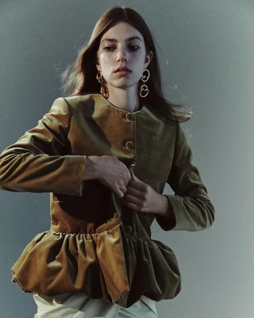 A young adult female buttons a short, fitted, light-brown velvet jacket in a studio setting with a grey background and soft, diffused lighting.