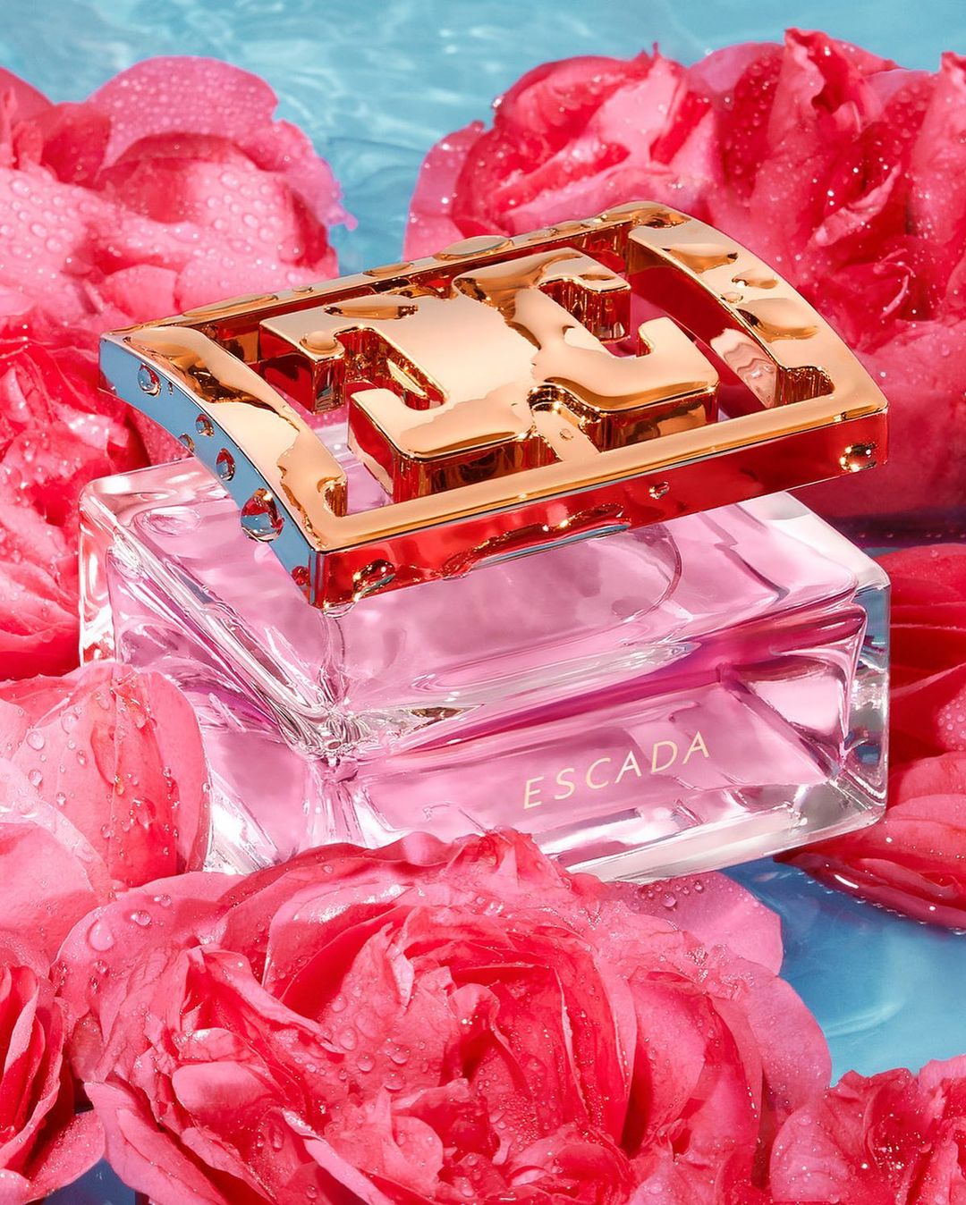 Escada perfume bottle sits atop a bed of pink roses with water droplets against a light blue background, creating a floral fragrance ad.