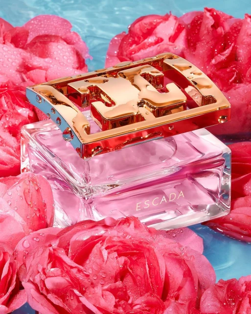 Escada perfume bottle sits atop a bed of pink roses with water droplets against a light blue background, creating a floral fragrance ad.