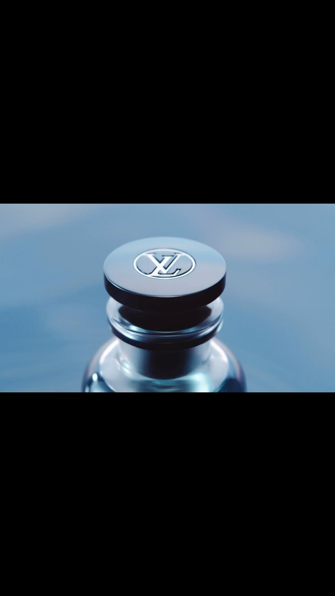 Close-up of a perfume bottle featuring the Louis Vuitton logo on its dark-colored cap against a blurred, dark blue background.