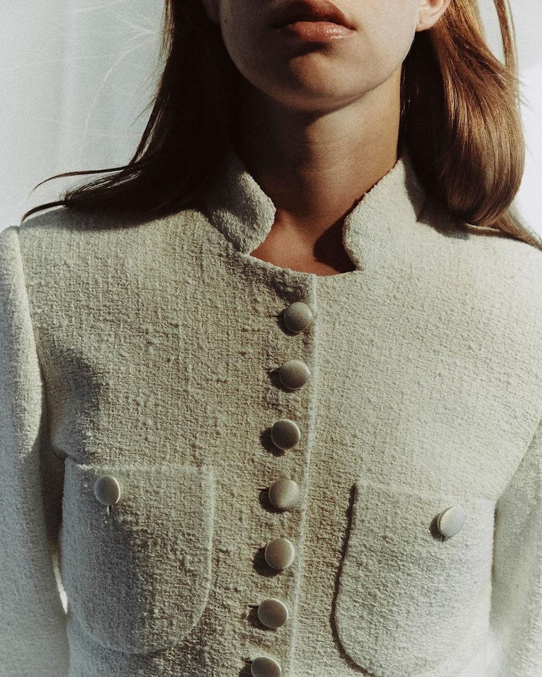 Close-up of a fair-skinned woman with light brown hair wearing a white tweed jacket with matching buttons, set against a white backdrop.