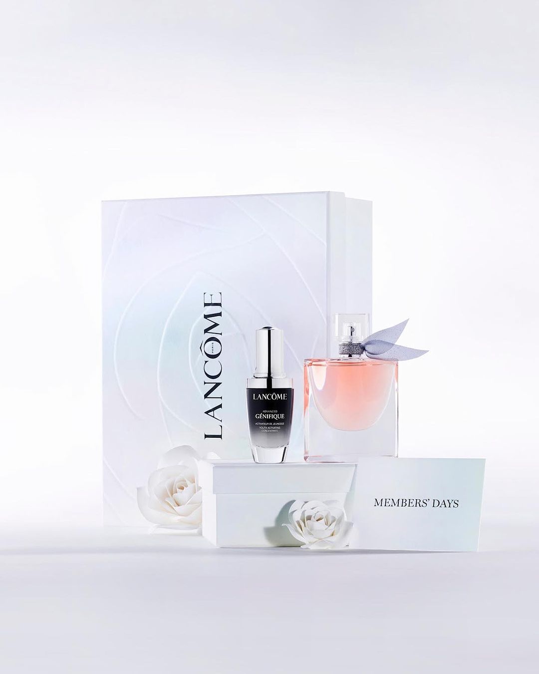 Product shot of Lancôme Genifique serum and La Vie Est Belle perfume with a branded gift bag and white roses on a white surface.