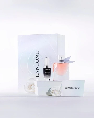 Product shot of Lancôme Genifique serum and La Vie Est Belle perfume with a branded gift bag and white roses on a white surface.