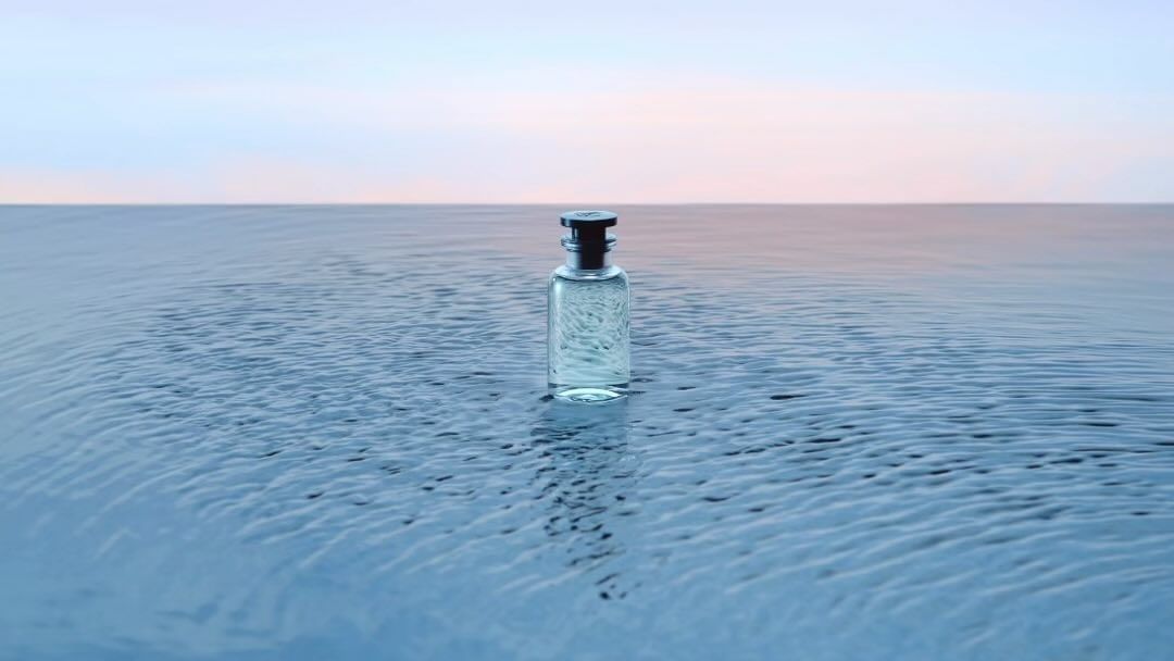 Clear glass perfume bottle with dark stopper stands centered on rippled water under a light pink and blue sky at the horizon.