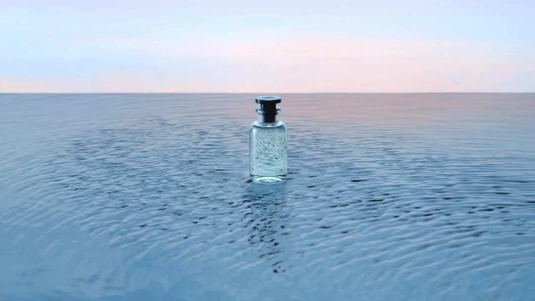 Clear glass perfume bottle with dark stopper stands centered on rippled water under a light pink and blue sky at the horizon.