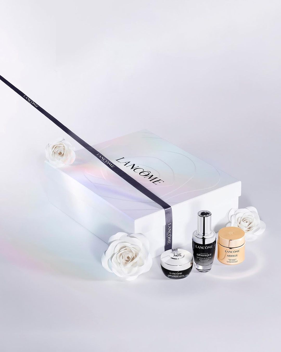 Lancôme beauty products displayed in front of a gift box with roses, sleek black ribbon and light color grading, product shot.