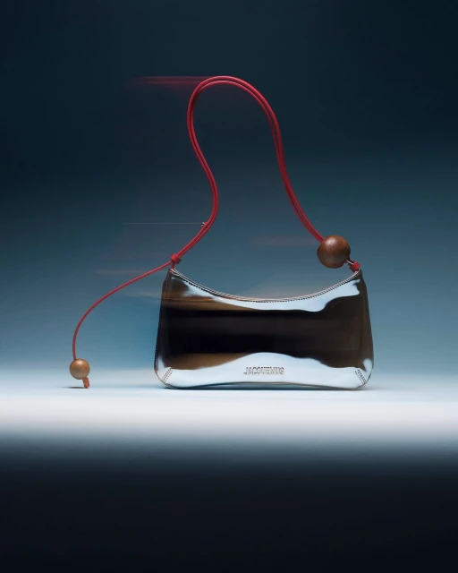 Designer handbag with silver reflective exterior, red shoulder strap, and wooden ball accents against a gradient blue background.