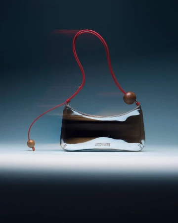Designer handbag with silver reflective exterior, red shoulder strap, and wooden ball accents against a gradient blue background.