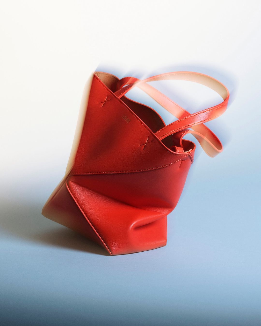 Studio shot of an orange leather tote bag with handles, against a soft blue and white background. Fashionable accessory close-up.