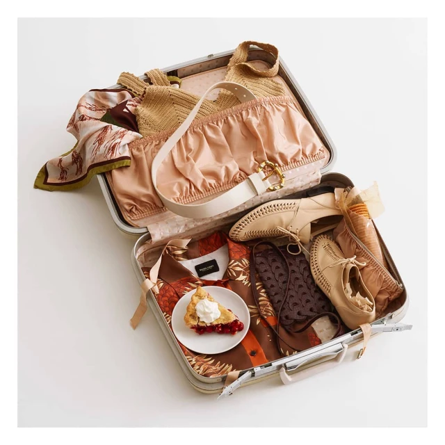 Overhead shot of a silver suitcase filled with clothing, shoes, and a piece of cherry pie on a white background, suggesting travel.