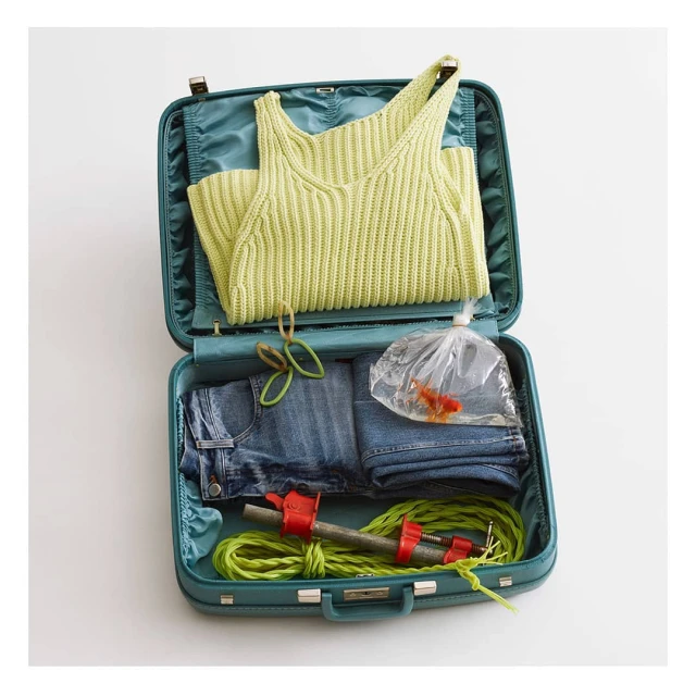 Open blue suitcase packed with clothing, rubber bands, workout equipment and a gold fish in a plastic bag on a white background.