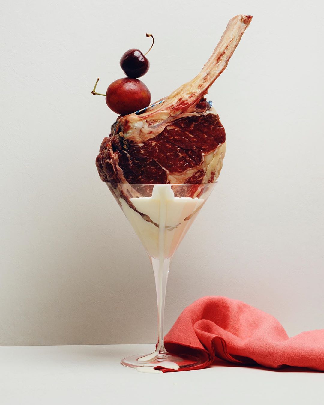 A bone-in ribeye steak served in a clear martini glass, topped with fresh cherries, and presented with a bright pink napkin.
