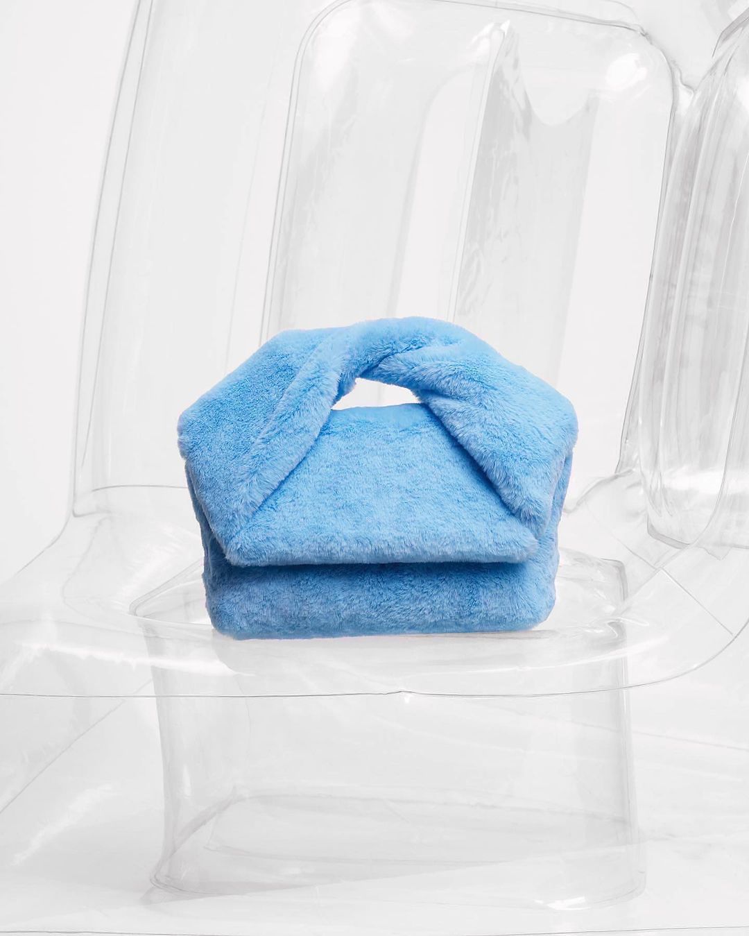 Light blue furry handbag with a twisted top handle sits on a clear inflatable chair against a white background in a studio setting.