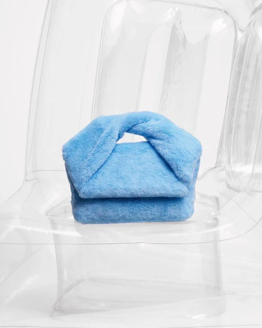 Light blue furry handbag with a twisted top handle sits on a clear inflatable chair against a white background in a studio setting.