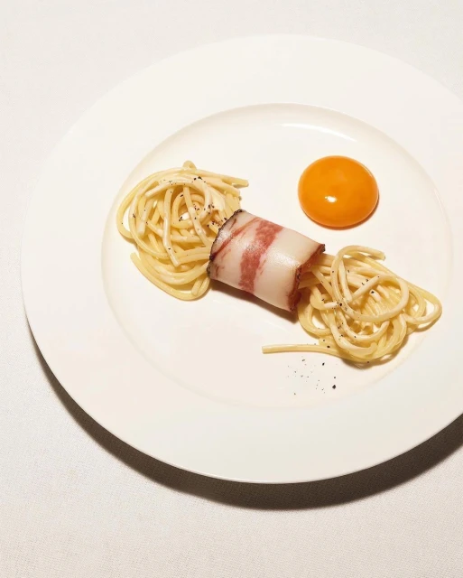 Overhead shot of spaghetti with an egg yolk and wrapped pancetta on a white plate against a white background, presented as fine dining.