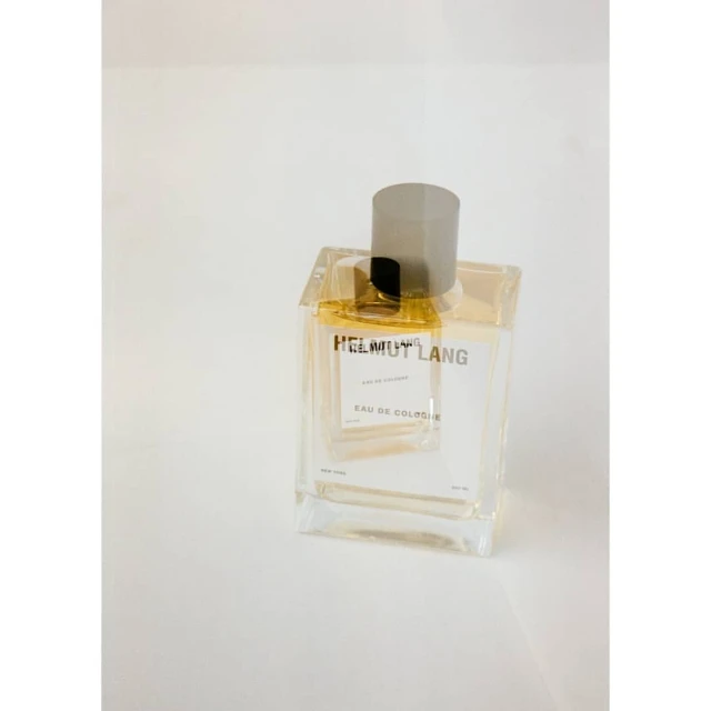 Still shot of clear glass HELMUT LANG Eau de Cologne bottle with silver top on a white surface. Perfume and luxury concept.