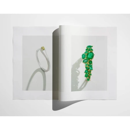Open magazine spread features a yellow and diamond bracelet plus an emerald and gold bracelet, jewelry advertisement, indoor shot.