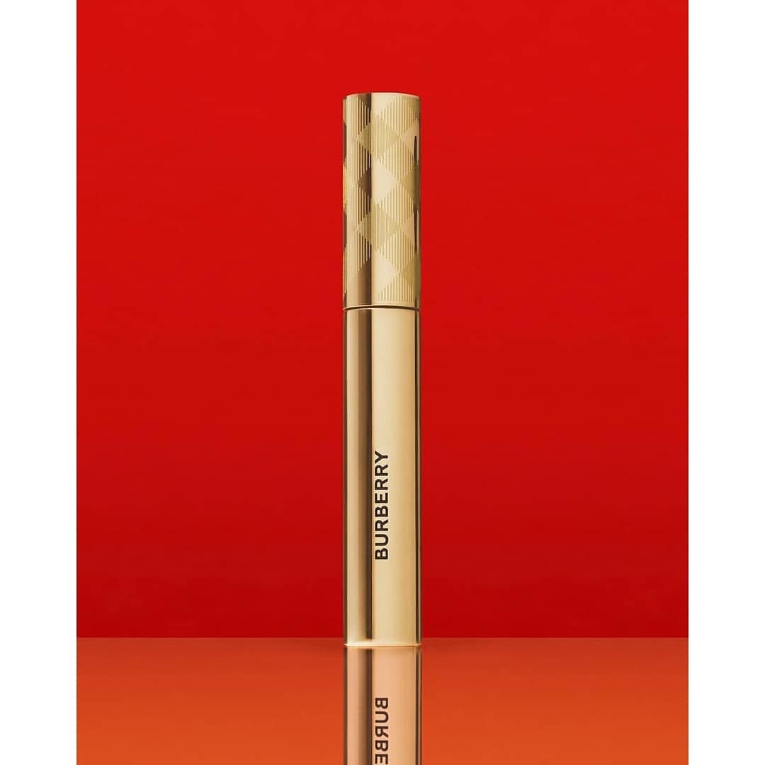 Close-up of a golden Burberry mascara tube standing upright against a vivid red background, showcasing luxury beauty product details.