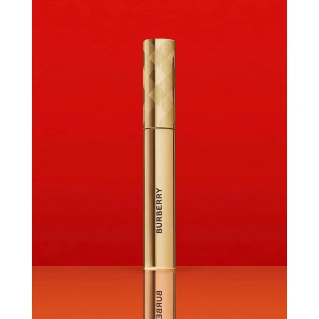 Close-up of a golden Burberry mascara tube standing upright against a vivid red background, showcasing luxury beauty product details.
