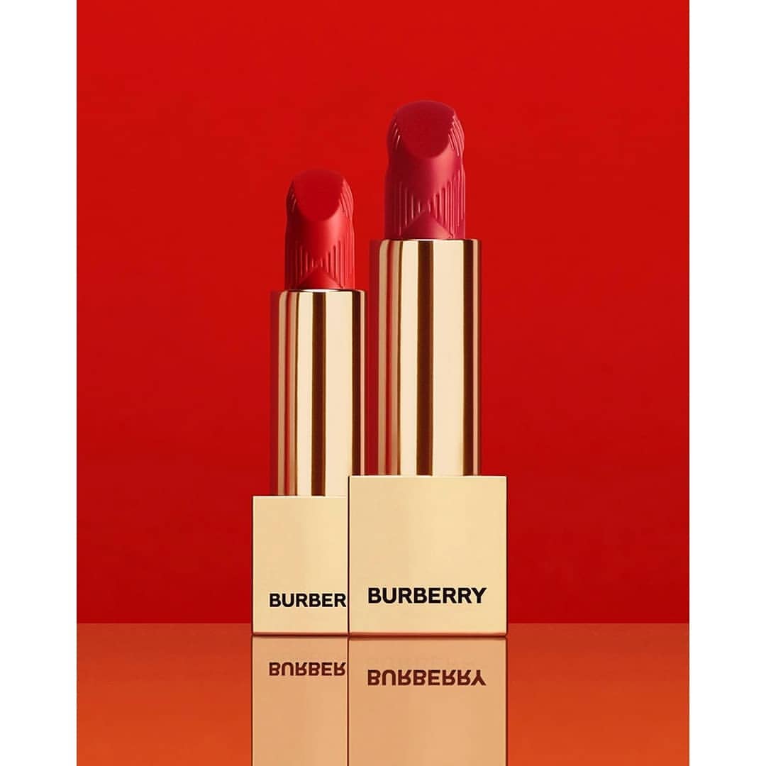 Two tubes of Burberry red lipstick stand upright on a reflective surface, with 'BURBERRY' printed on the base against a red backdrop.