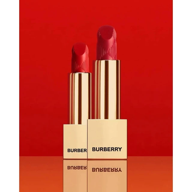Two tubes of Burberry red lipstick stand upright on a reflective surface, with 'BURBERRY' printed on the base against a red backdrop.