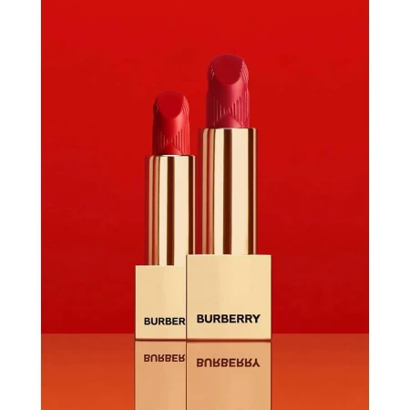 Two tubes of Burberry red lipstick stand upright on a reflective surface, with 'BURBERRY' printed on the base against a red backdrop.