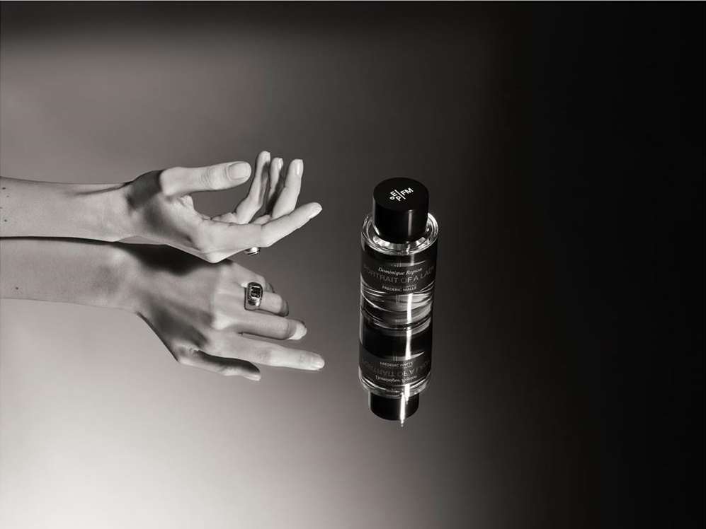 Elegant perfume bottle presented with woman's hands adorned with rings against a smooth, contrasting black and gray background in a product shot.