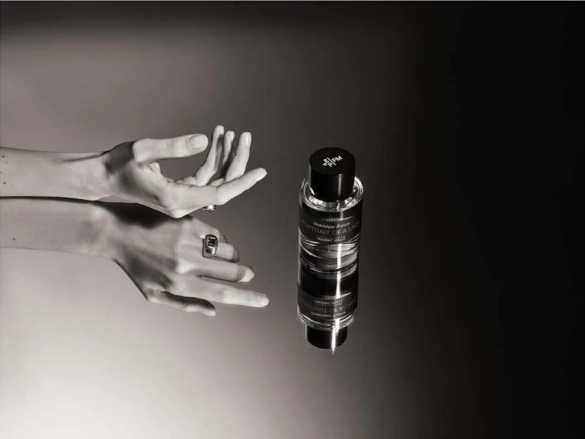 Elegant perfume bottle presented with woman's hands adorned with rings against a smooth, contrasting black and gray background in a product shot.