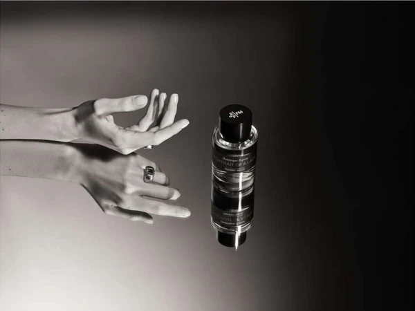 Elegant perfume bottle presented with woman's hands adorned with rings against a smooth, contrasting black and gray background in a product shot.