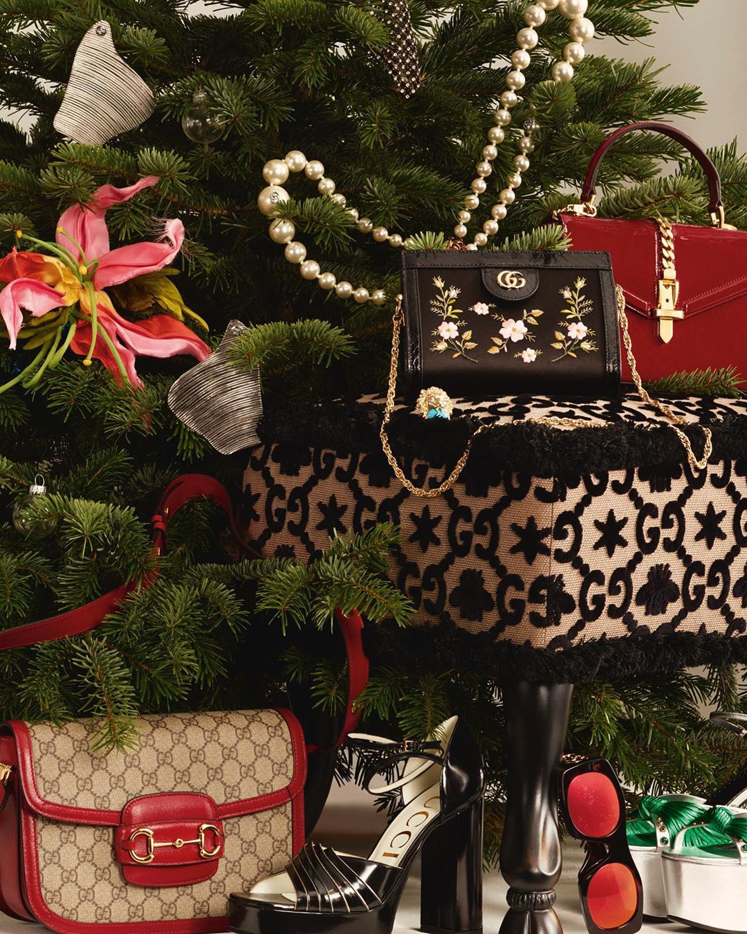 Luxury Christmas still life featuring Gucci handbags and high-heeled platform shoes arranged around a decorated Christmas tree with ornaments.