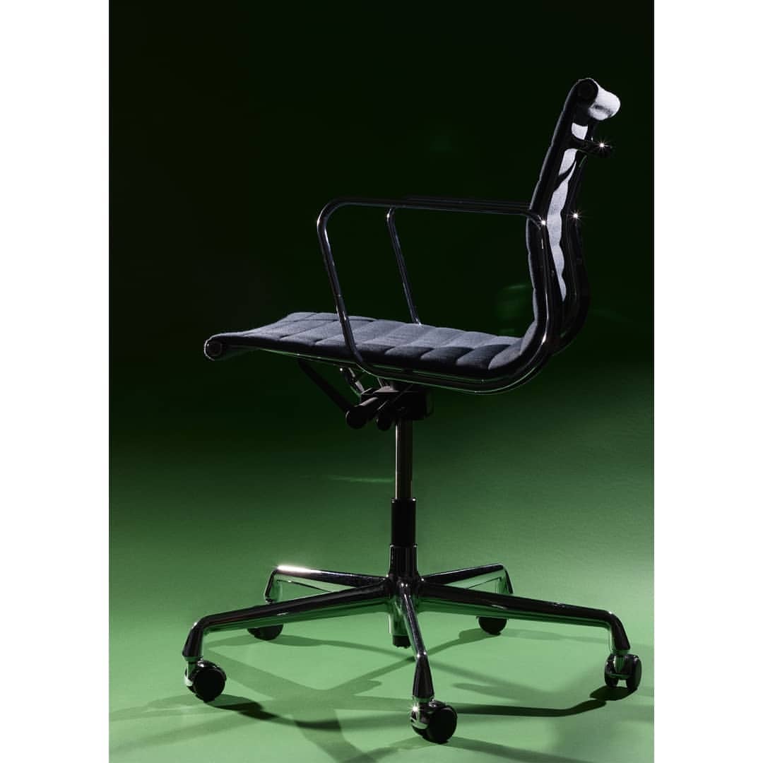 A black office chair with a chrome frame is positioned against a green background in a well-lit interior space, highlighting its design.
