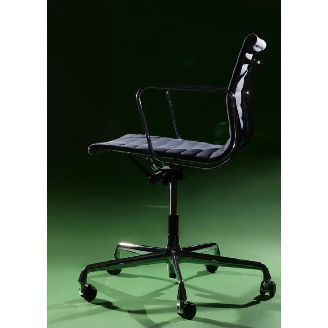A black office chair with a chrome frame is positioned against a green background in a well-lit interior space, highlighting its design.