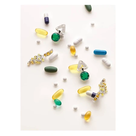 Still life studio shot of green gemstone jewelry with diamonds among assorted pharmaceutical capsules and pills scattered on a white surface.