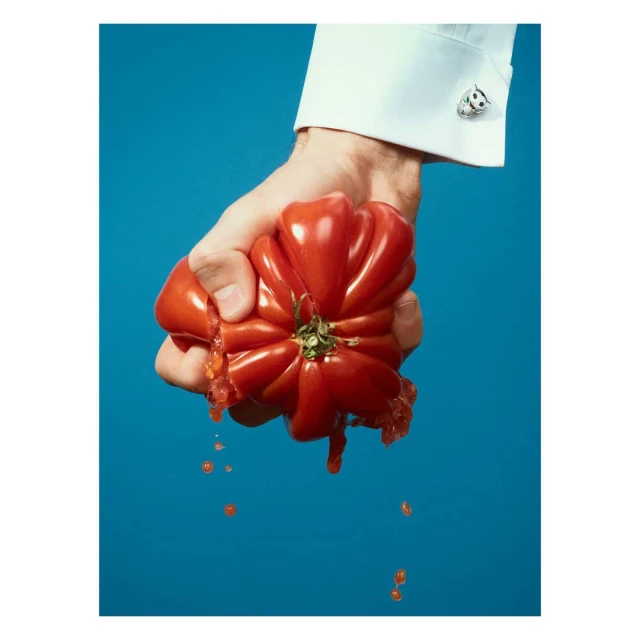 Close-up of a hand squeezing a large tomato, with juice dripping down, set against a bright blue background, showcasing white shirt and cufflink.