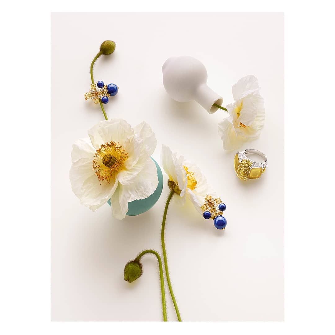 Still life shot of white poppy flowers in vases with jewelry including a yellow diamond ring, studio setting against a white background.