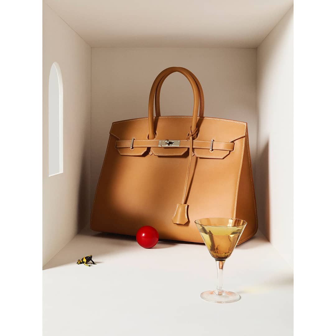 Tan Birkin bag in a white niche with a red sphere and a martini glass filled with a yellow liquid on a white surface.