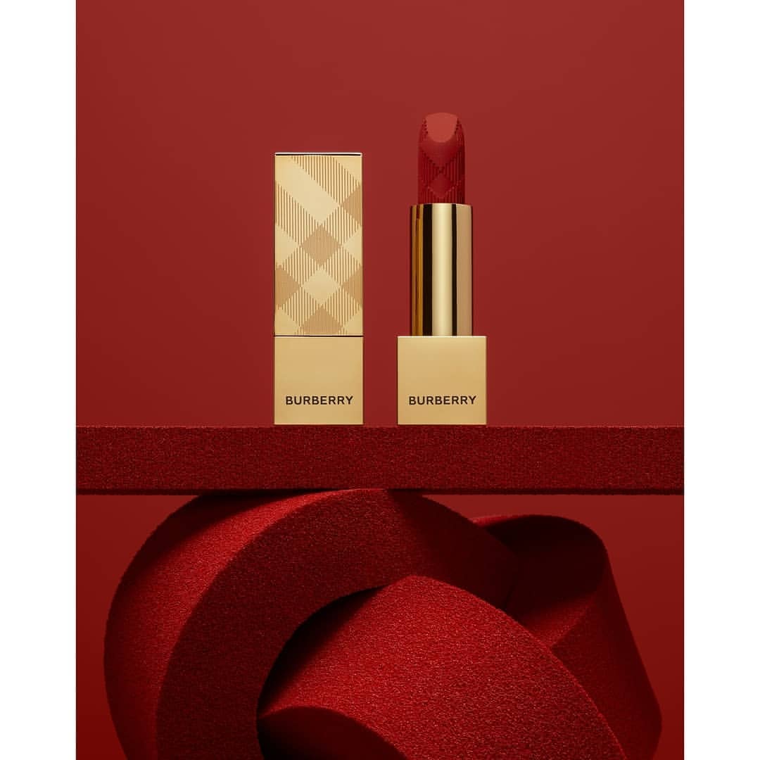Close-up studio shot of gold Burberry lipstick with red lipstick, standing on a red textured surface against a solid red background.