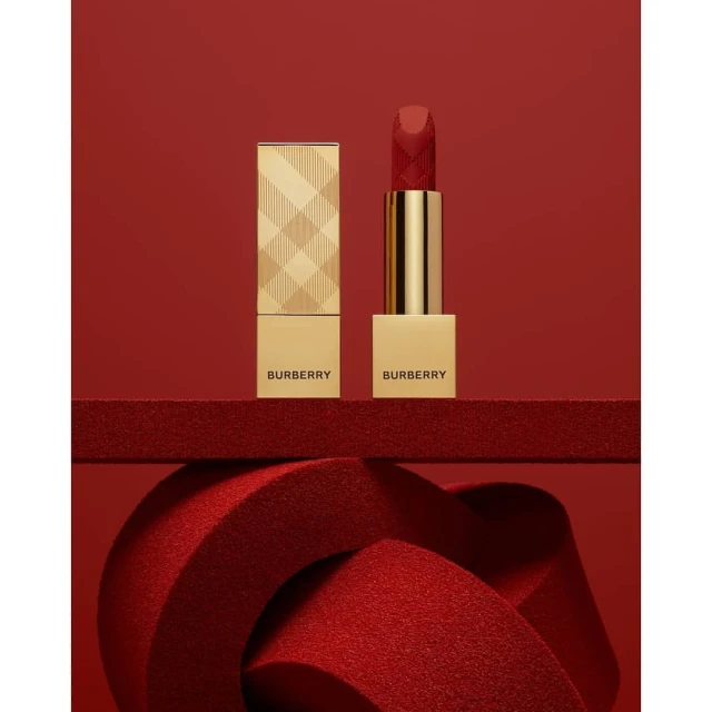 Close-up studio shot of gold Burberry lipstick with red lipstick, standing on a red textured surface against a solid red background.