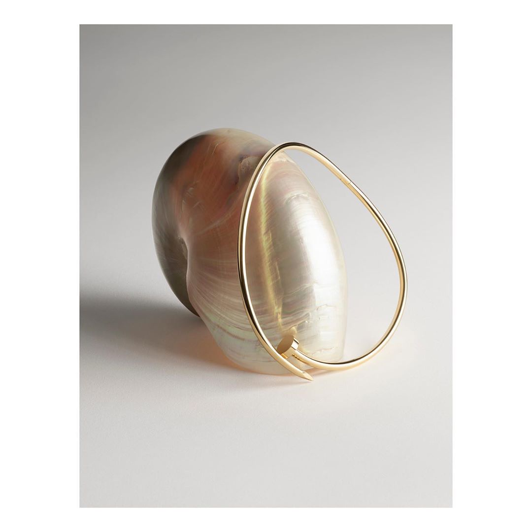 Gold bracelet rests against a large iridescent seashell on a pale grey background, creating an elegant and minimalist composition.