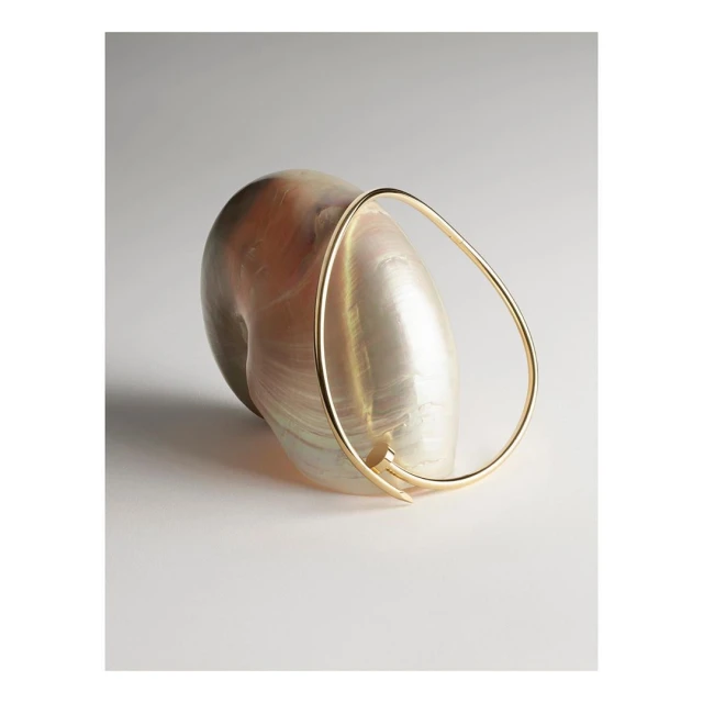 Gold bracelet rests against a large iridescent seashell on a pale grey background, creating an elegant and minimalist composition.