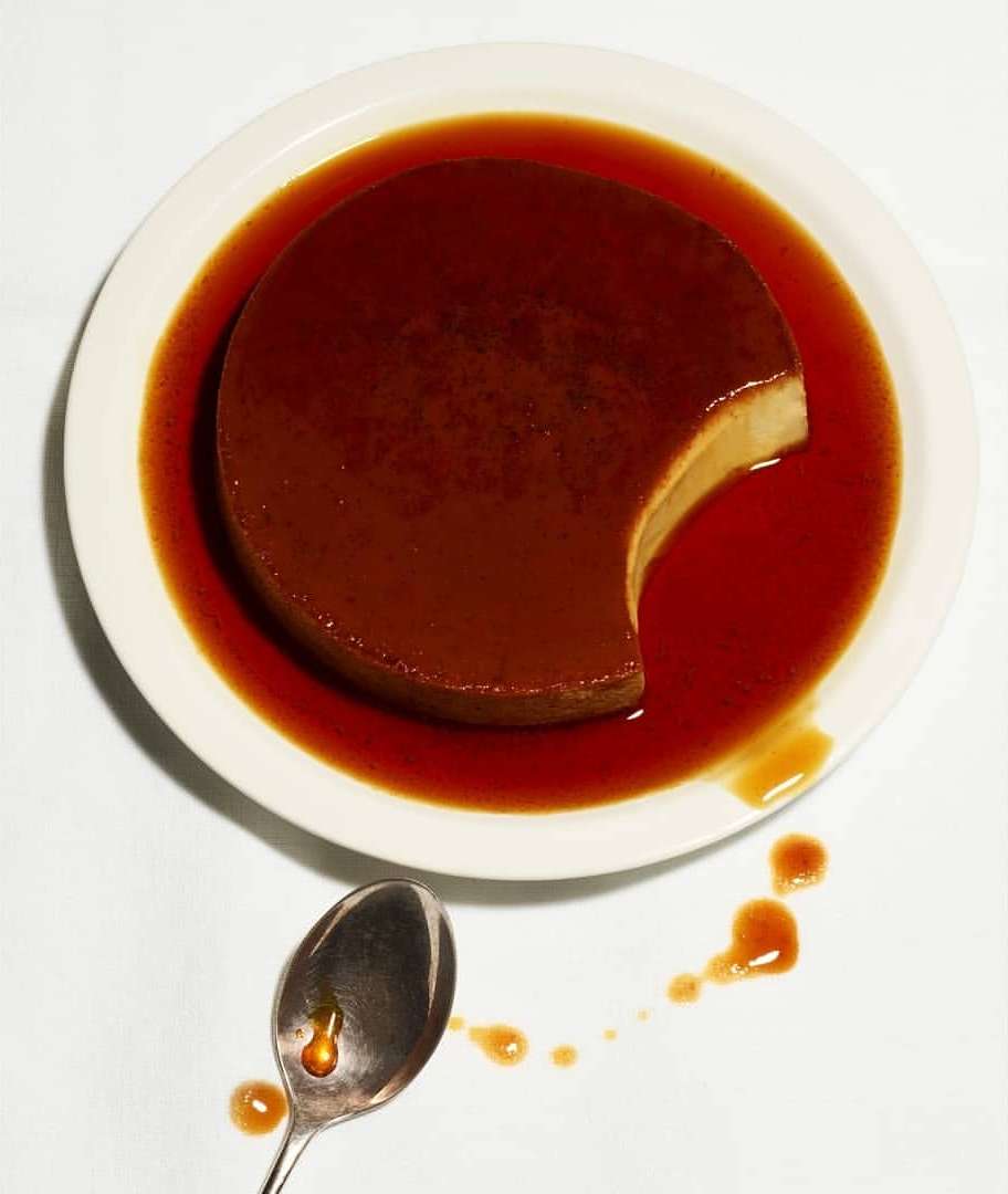 Close-up shot of a caramel flan dessert with a bite taken out, served on a white plate with a silver spoon and caramel sauce.