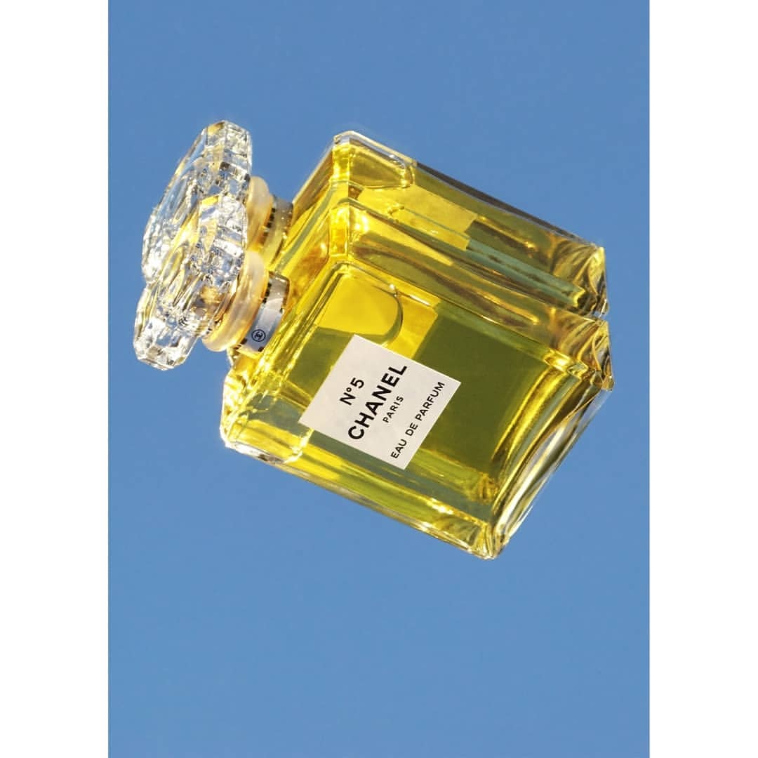Crystal-topped Chanel No. 5 perfume bottle containing yellow liquid on a blue reflective surface. The bottle is tilted and shows its label.