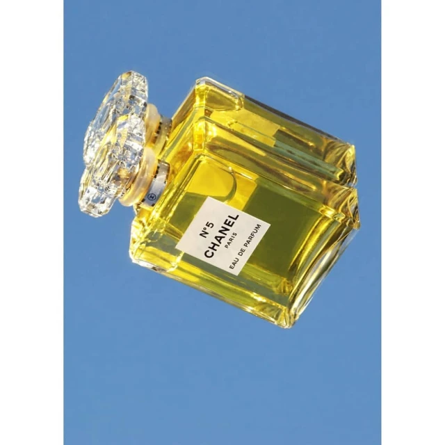 Crystal-topped Chanel No. 5 perfume bottle containing yellow liquid on a blue reflective surface. The bottle is tilted and shows its label.
