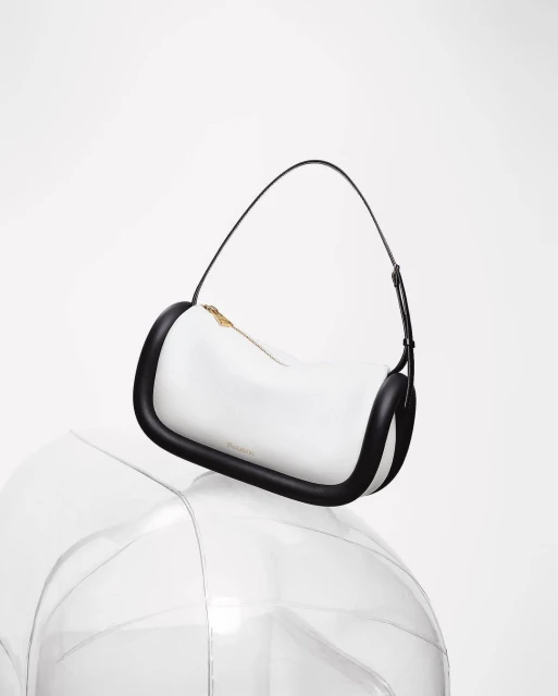 Fashion shot of a white handbag with black trim and black handle, resting on a clear, transparent sphere in a white photo studio.