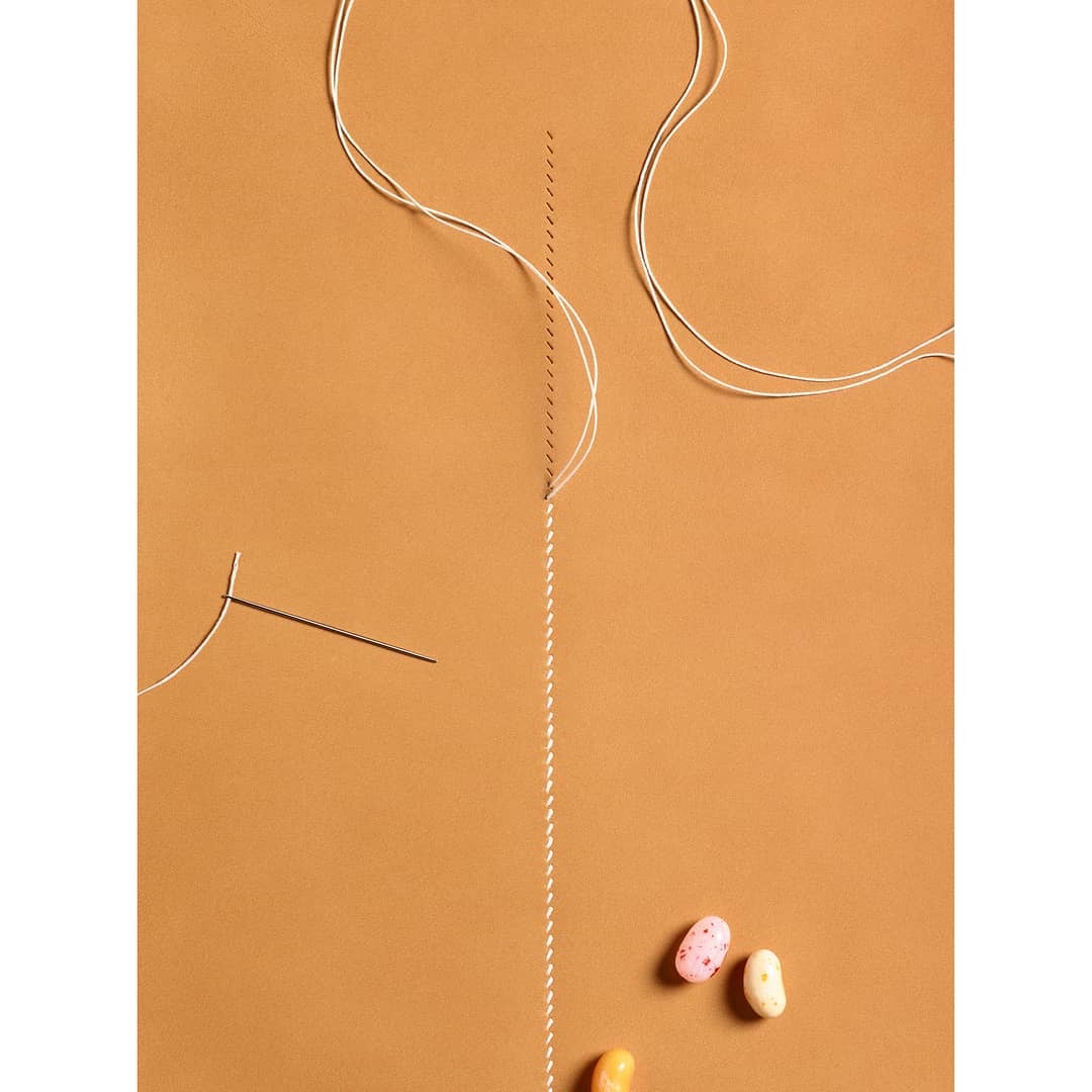 Overhead shot showcasing a sewing needle, white thread, and colorful jelly beans on a solid tan background, creatively arranged.