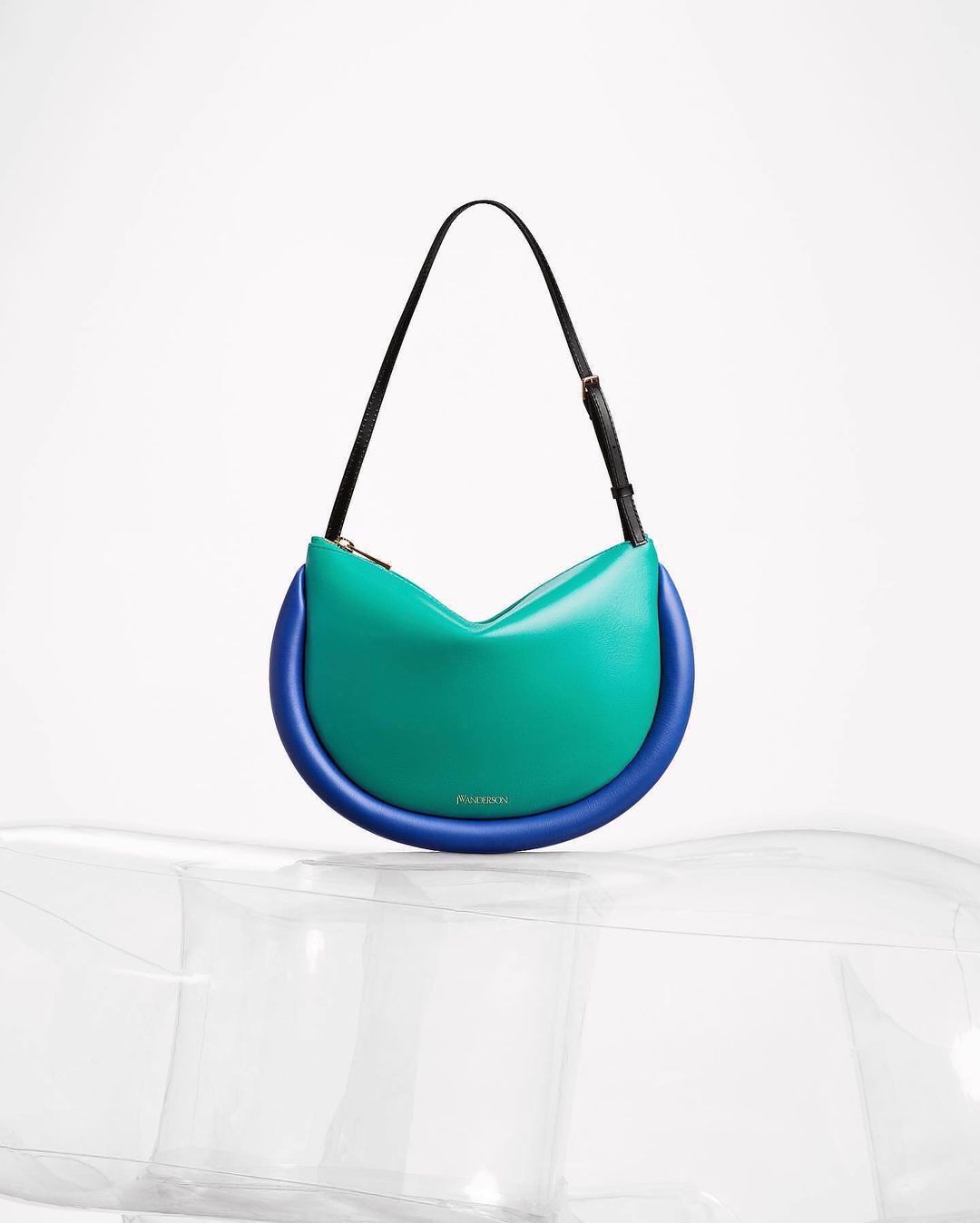 Fashion product shot of a JW Anderson Bumper-15 bag in turquoise and blue on a clear, inflatable display stand against white.