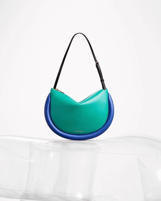 Fashion product shot of a JW Anderson Bumper-15 bag in turquoise and blue on a clear, inflatable display stand against white.